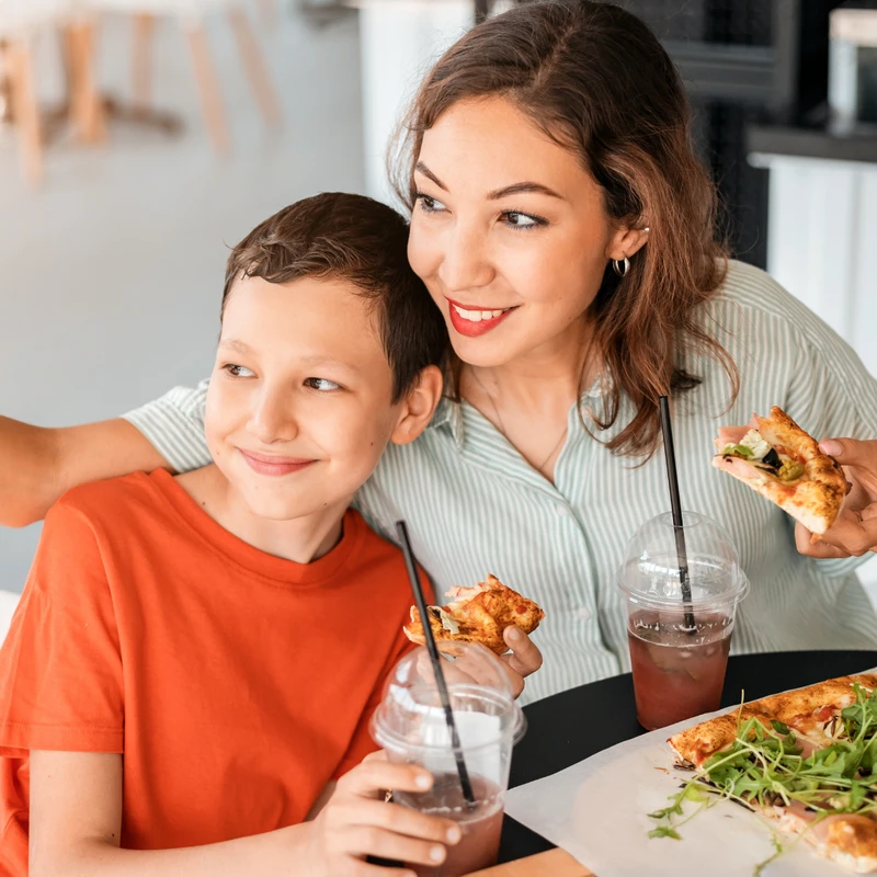A smiling mother and her son sit at a restaurant table, taking a selfie with a smartphone while eating pizza and holding drinks.