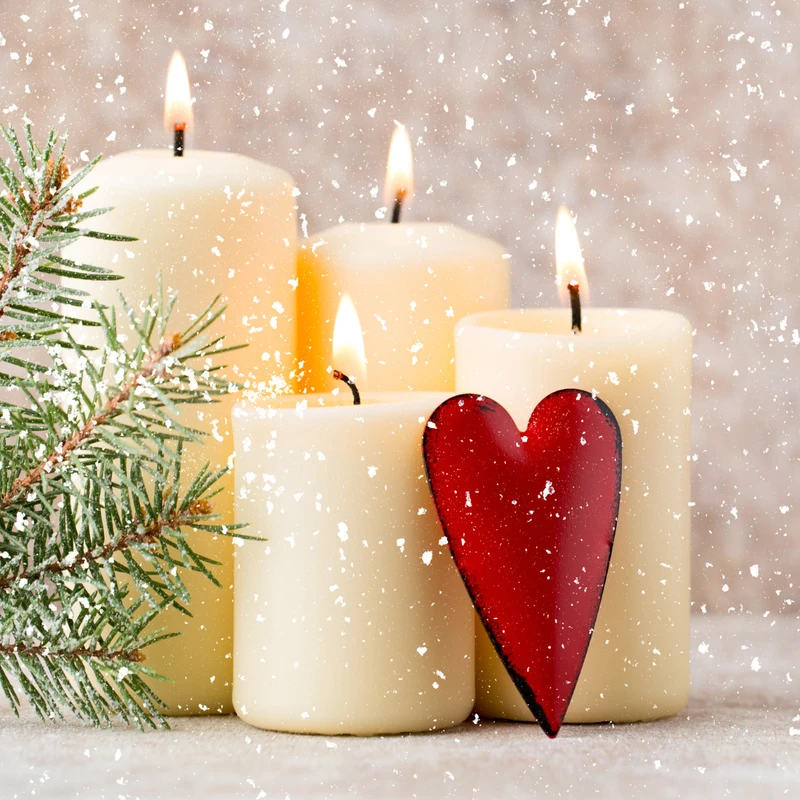 Four lit white pillar candles sit next to a snow-dusted pine sprig and a red heart ornament, with artificial snow falling all around them.
