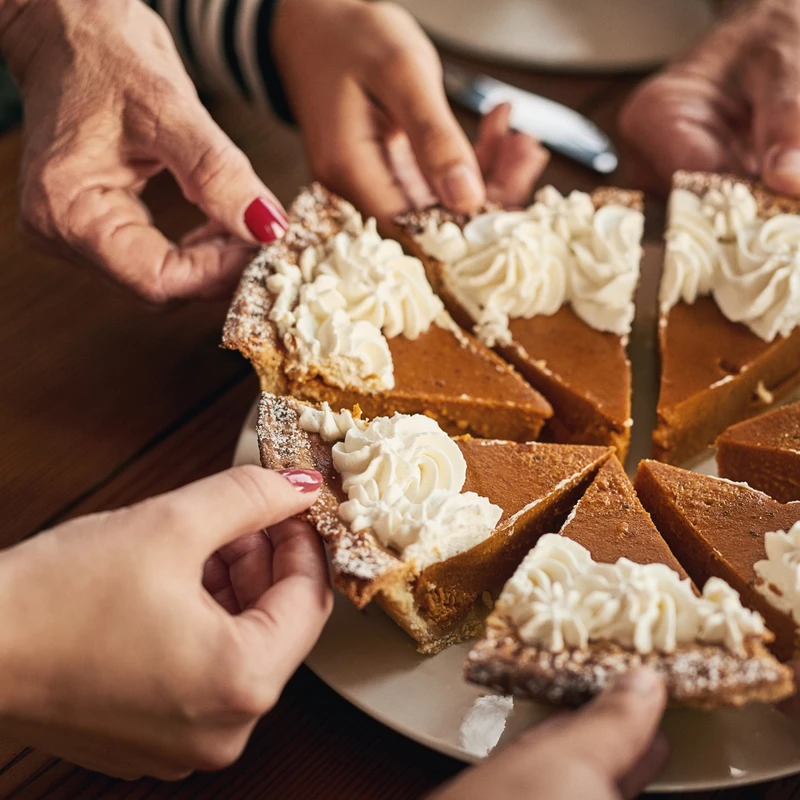 Many hands reach in to take slices of a pumpkin pie topped with whipped cream from a plate on a wooden table, suggesting a family gathering.