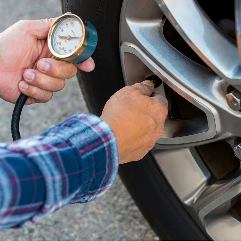A person wearing a plaid shirt uses a tire pressure gauge to check the air in a car's tire with a silver alloy wheel.