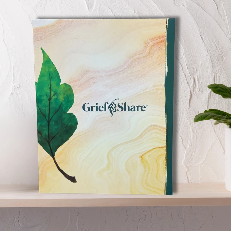 The GriefShare workbook stands on a light wooden shelf next to a white pot of yellow flowers, against a textured white wall.