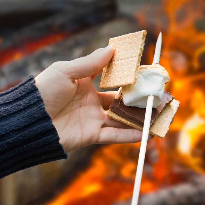 A person's hand in a sweater assembles a s'more with a toasted marshmallow on a stick in front of a glowing campfire.