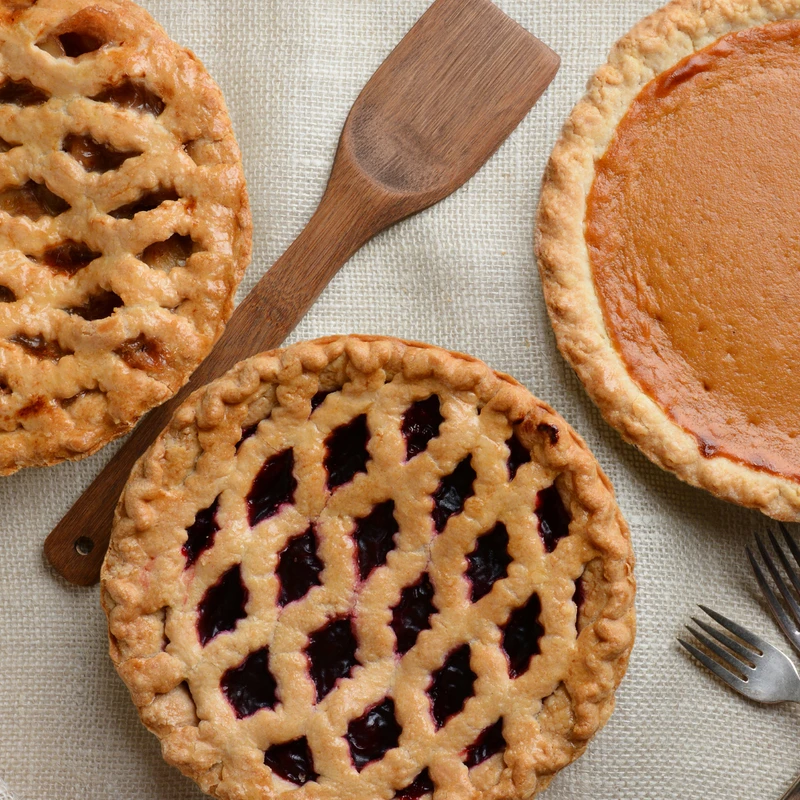An overhead view of three pies on a burlap cloth: a lattice-topped apple pie, a lattice-topped berry pie, and a pumpkin pie, with forks and a spatula.