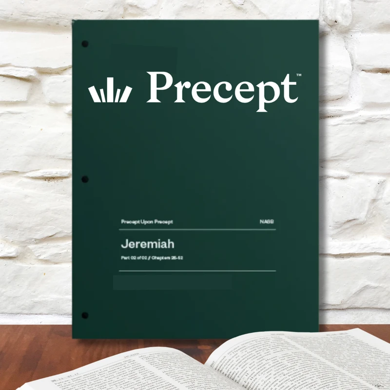 A dark green Precept Bible study guide for Jeremiah stands on a wooden desk behind an open book, with a white stone wall in the background.