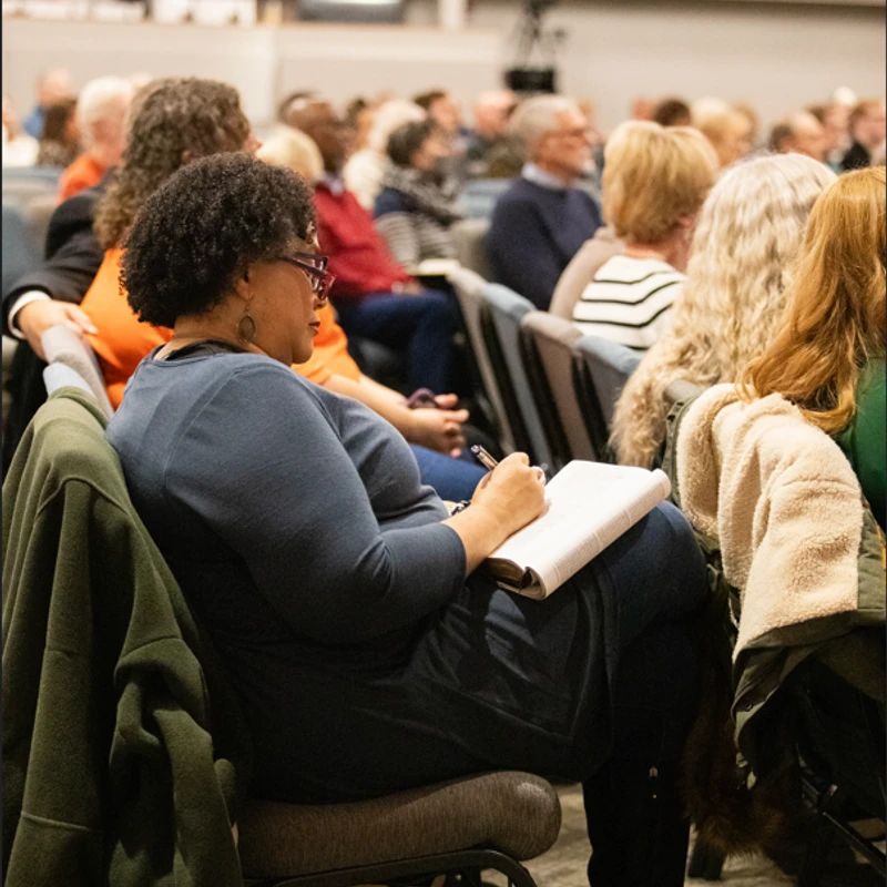 A Black woman with curly hair and glasses sits in an audience, focused on taking notes in a book on her lap during a presentation.