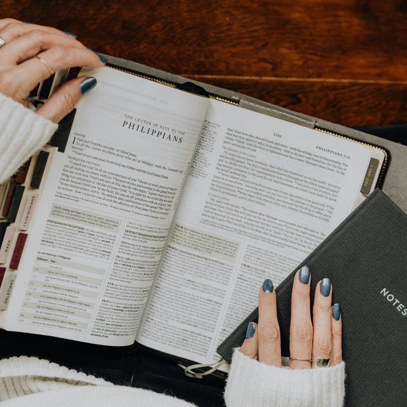 An overhead shot of a person wearing a white sweater reading a Bible open to the book of Philippians, with a notebook resting on their lap.