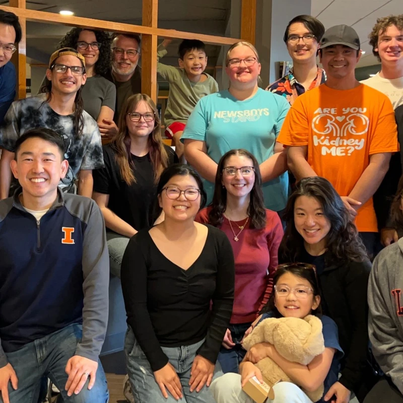 A large, diverse group of about 17 smiling people of various ages gathered together indoors for a happy group portrait.