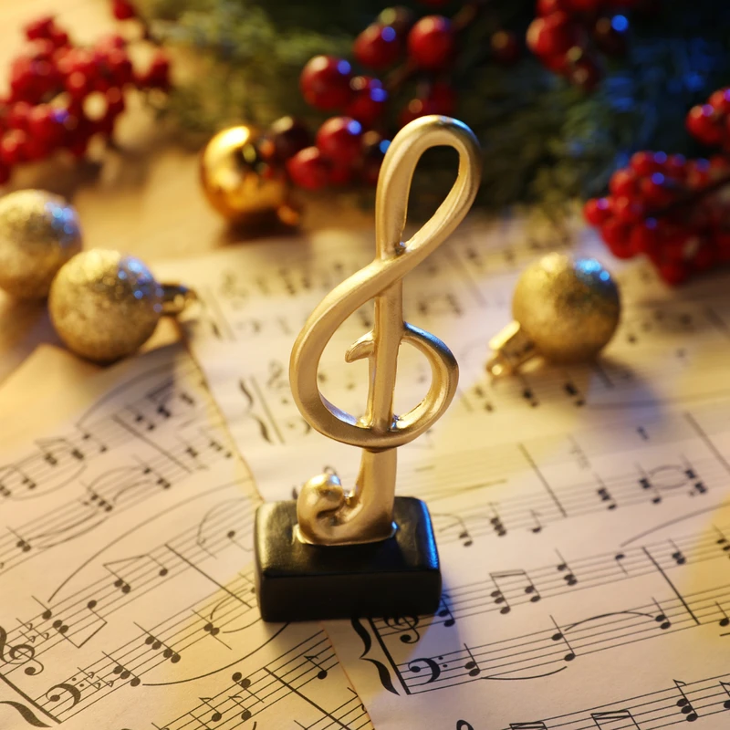 A golden treble clef statue stands on sheet music, surrounded by gold Christmas ornaments, red berries, and pine branches on a wooden table.