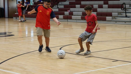 Two young boys in red shirts play with a soccer ball on a polished wooden gymnasium floor, with bleachers and signs for the 'Rams' in the background.