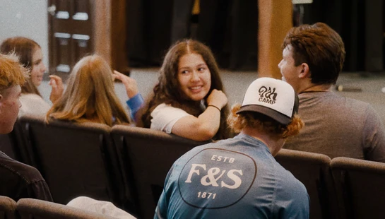 A group of young people are sitting in rows of chairs in an auditorium. One girl is turned around smiling and talking to the people behind her.