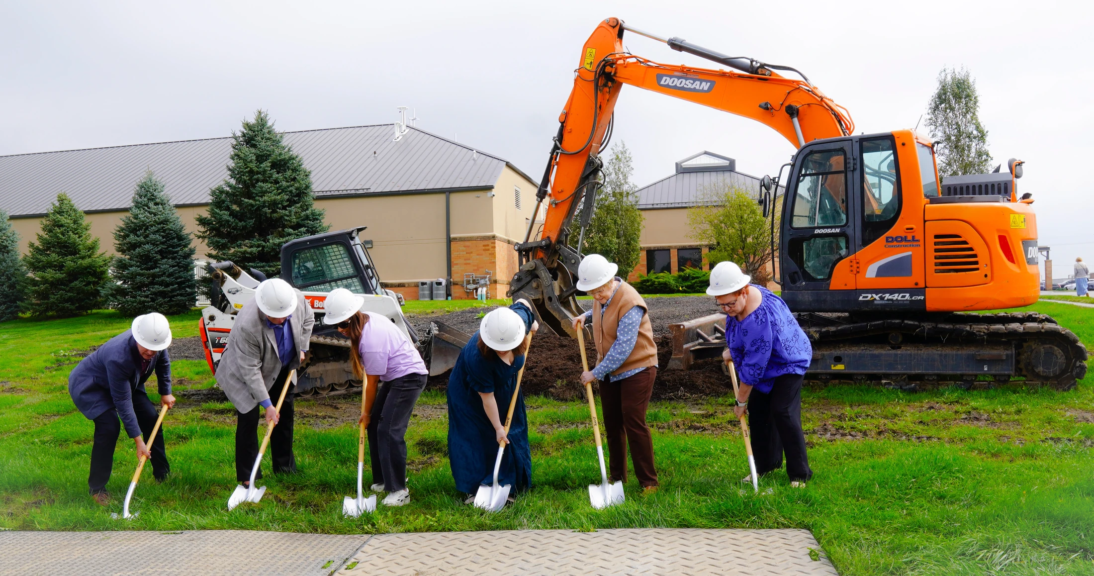 A group of six people in white hard hats use shovels to break ground on a grassy field, with a large orange excavator behind them.
