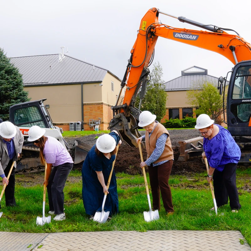 A group of six people in white hard hats use shovels to break ground on a grassy field, with a large orange excavator behind them.