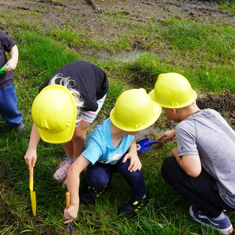 An overhead view of four children wearing yellow hard hats, playing and digging in a muddy, grassy area with small toy shovels.
