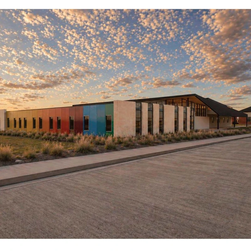 A long, modern building with a rainbow-colored facade sits in a flat field under a beautiful sunset sky filled with small, puffy clouds.