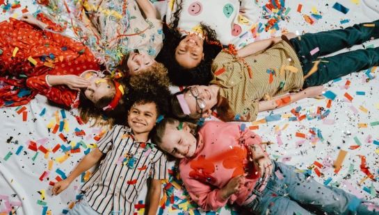 A top-down view of a diverse group of happy children lying in a circle on the floor, surrounded by and showered in colorful confetti.