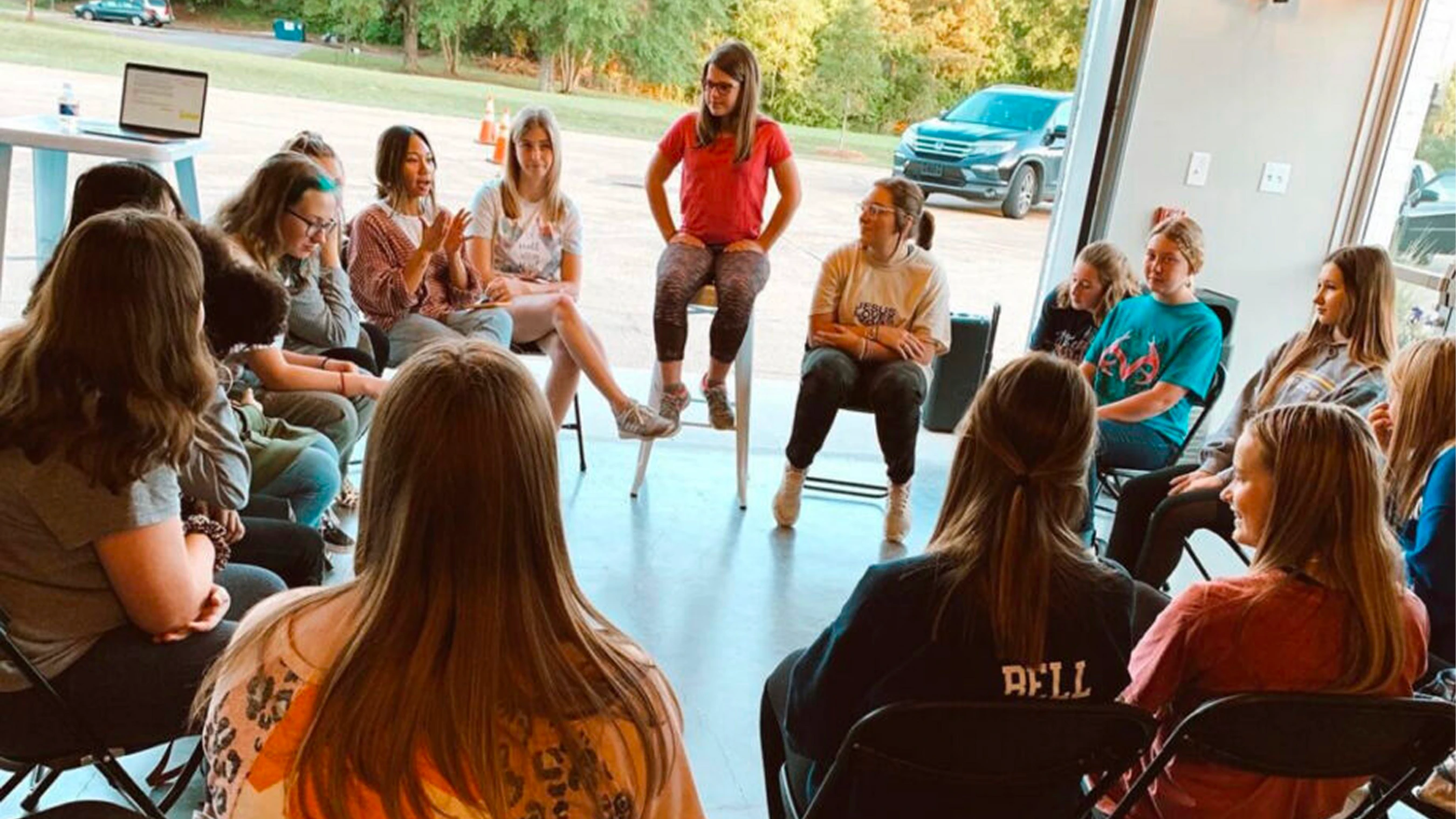 A group of teenage girls sits in a circle of chairs, having a discussion. One girl in a red shirt sits on a stool in the center, leading the conversation.