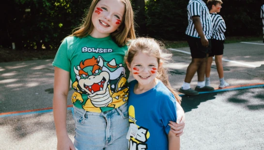 Two young girls with red face paint on their cheeks smile at the camera. The older girl wears a green Bowser shirt and hugs the younger girl in a blue shirt.
