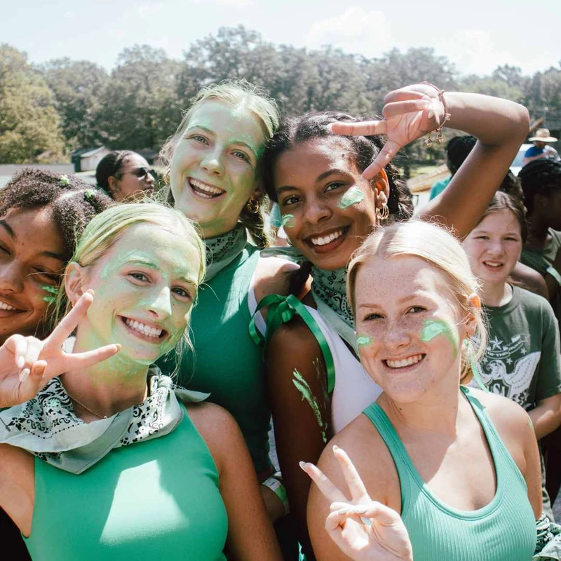 A group of happy young women dressed in green, with green face paint on, smile and make peace signs for the camera on a sunny day outdoors.