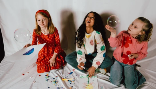 Three diverse young girls sit on a white sheet, happily drawing, painting, and playing with soap bubbles surrounded by colorful art supplies.