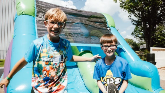 Two young boys smile for the camera while standing on a large, colorful inflatable slide outdoors on a sunny day.