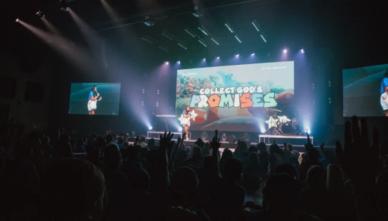 View from the audience of a person on a brightly lit stage. A large screen reads "Collect God's Promises," and many people in the crowd have their hands raised.