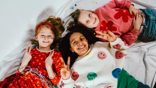 An overhead shot of three diverse young girls lying on a white sheet. They are smiling, making peace signs, and have confetti and stickers on their faces.