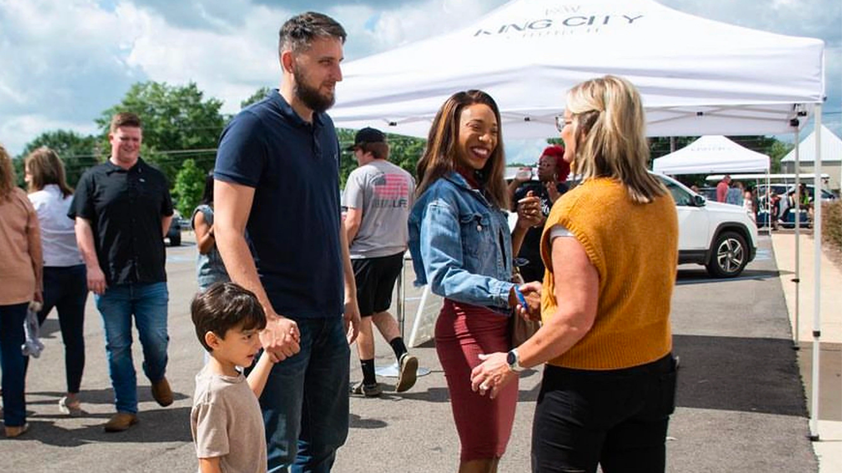 A group of people socializing outdoors. A man holds a boy's hand while two women smile and shake hands in front of a white event tent.