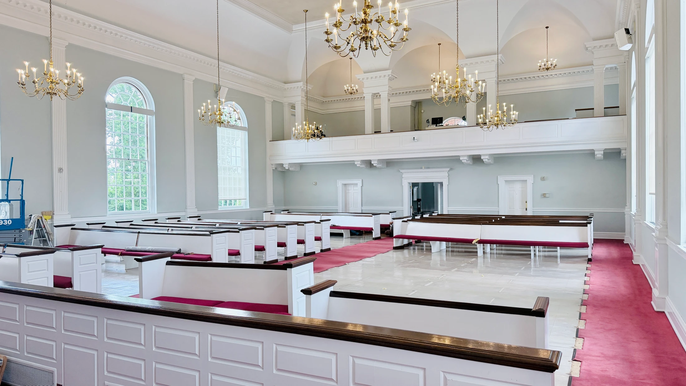 A bright, airy church sanctuary with white pews, red carpeting, and ornate chandeliers is shown during renovation, with a construction lift in the corner.