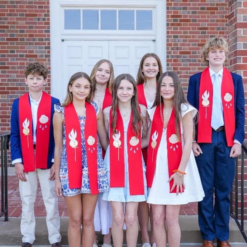 A group of seven teenagers, five girls and two boys, stand on the steps of a brick church wearing red stoles for their confirmation ceremony.