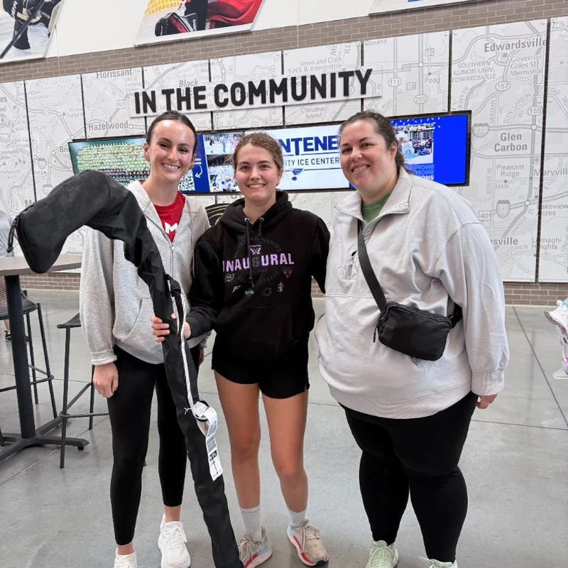 Three smiling women stand side-by-side for a photo in front of a wall that reads 'In the Community' with graphics of hockey players at an ice center.