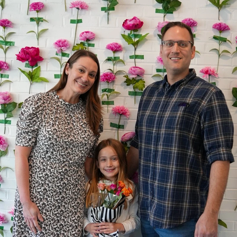A smiling family of three poses for a photo. The mother, father, and young daughter stand in front of a white brick wall decorated with pink flowers.