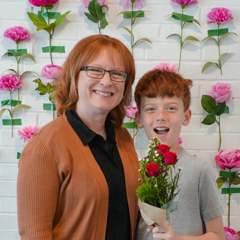 A smiling woman with red hair and glasses stands beside a happy young boy holding a small bouquet of red roses against a white brick wall with pink flowers.