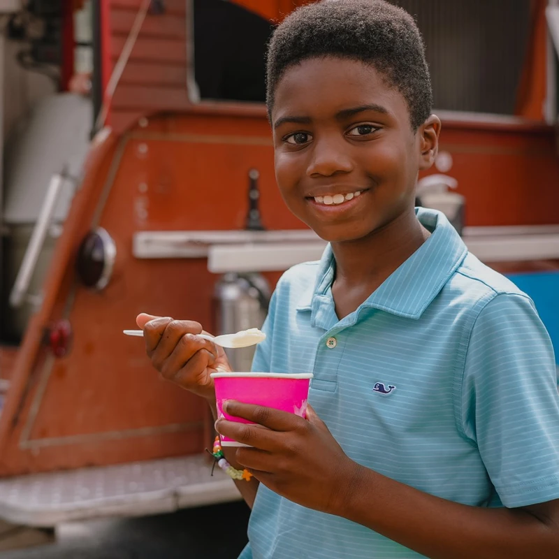 A young Black boy in a blue striped polo shirt smiles while holding a pink cup and a spoon, eating ice cream in front of a red food truck.