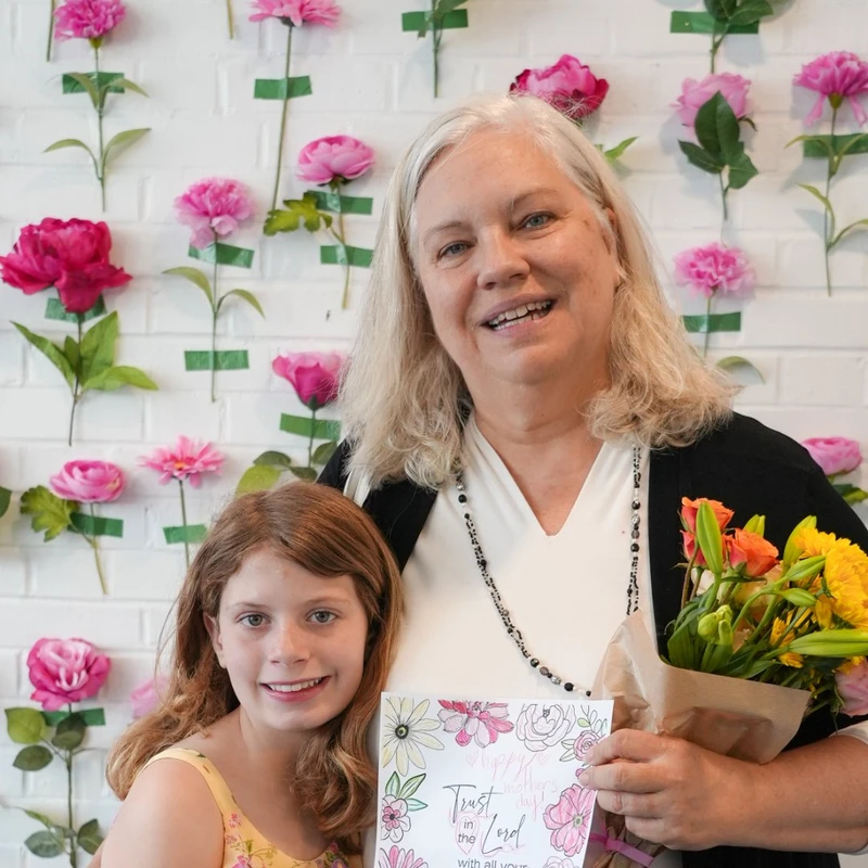 A smiling older woman and a young girl pose together. The woman holds a bouquet of flowers and a card, standing in front of a white wall with flowers.