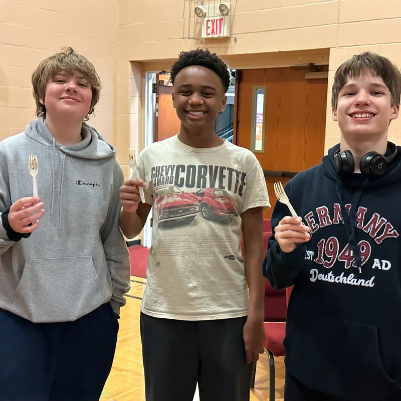 Three smiling teenage boys stand side-by-side indoors. Each boy is holding up a white plastic fork, posing for the camera at what appears to be a gathering.