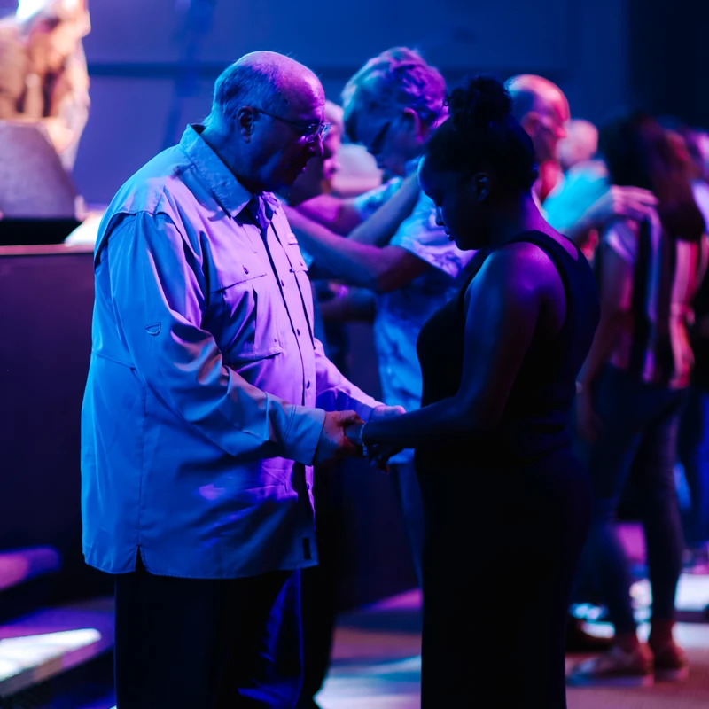 An older man and a younger woman hold hands and bow their heads in prayer during a church service, illuminated by blue and purple stage lights.