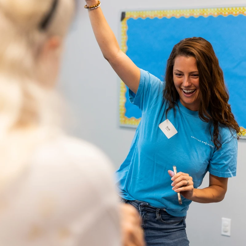 A woman with long brown hair smiles broadly as she raises her hand in the air. She wears a blue t-shirt and jeans and holds a phone in her other hand.