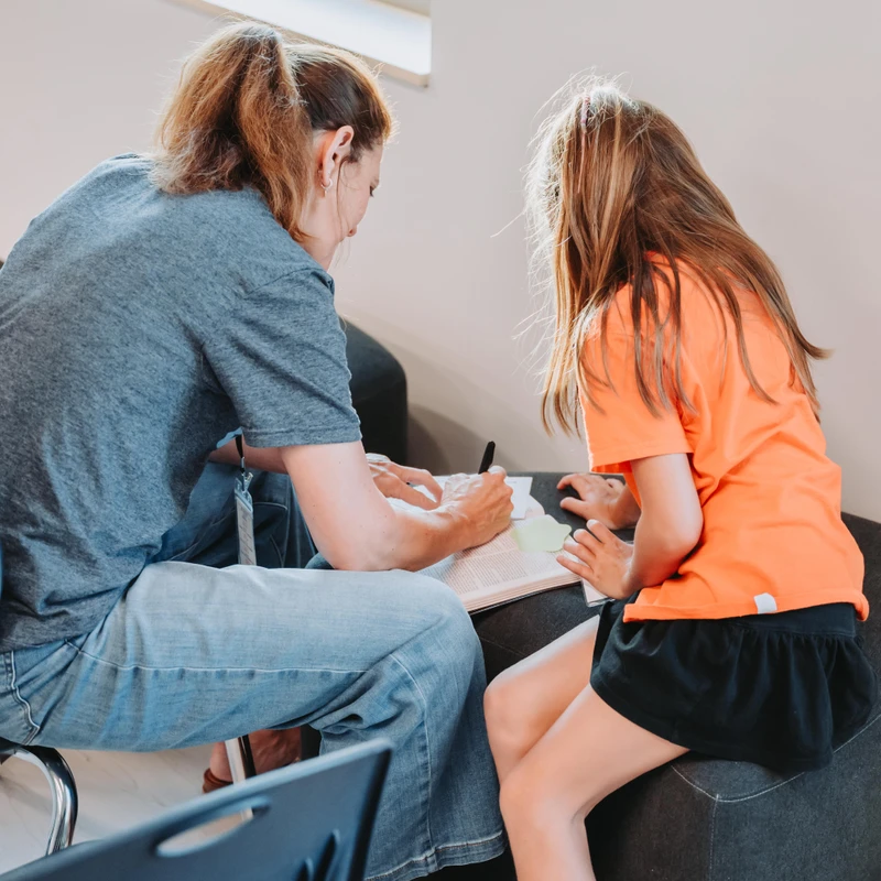 A woman and a young girl sit next to each other, focused on a book open on their laps. The woman is writing in the book, seemingly teaching the girl.