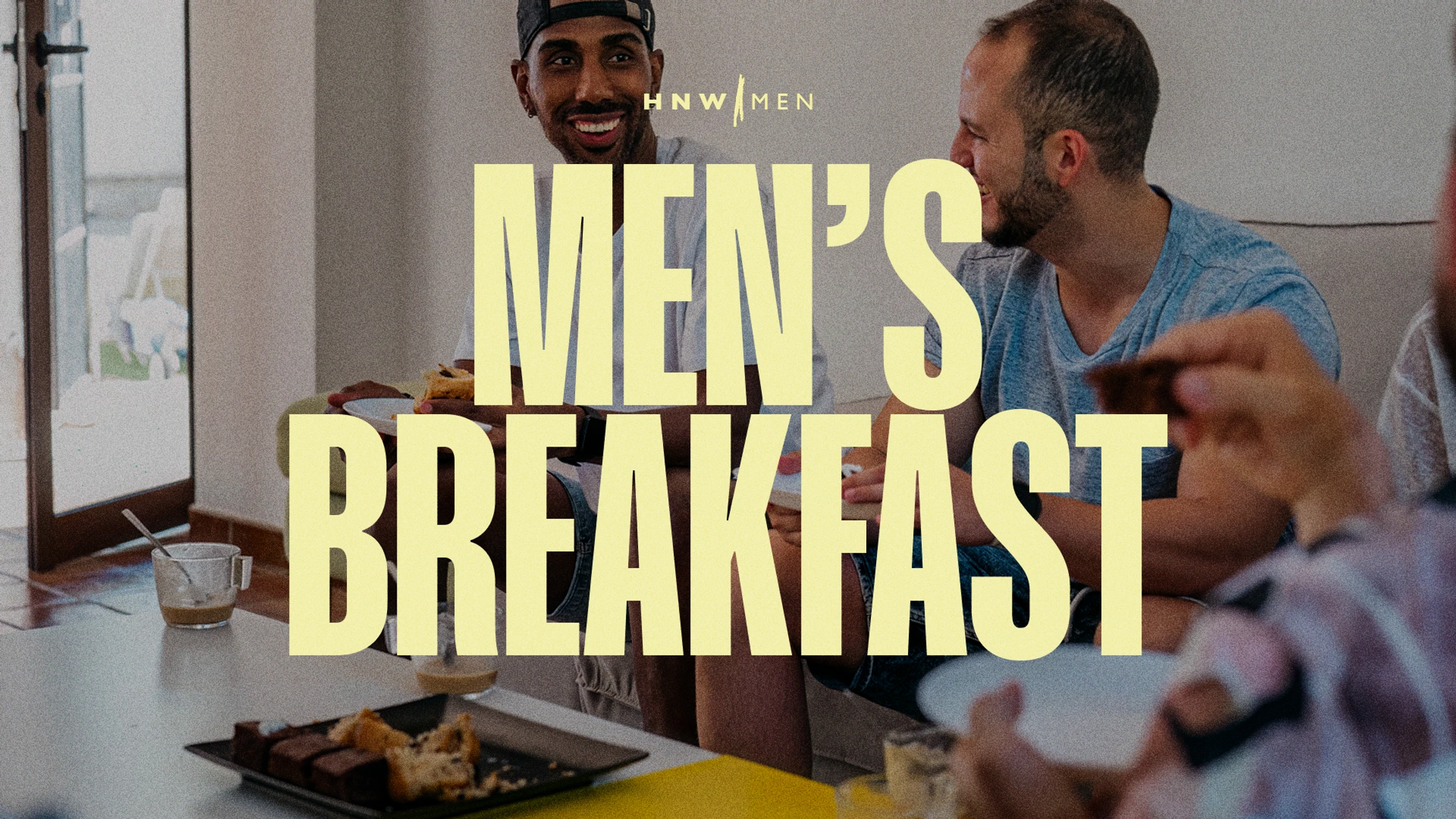 A diverse group of men smile and socialize while eating together at a table. Bold yellow text over the image reads "Men's Breakfast".