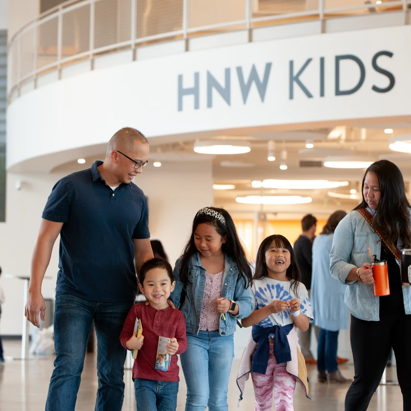 A family of five with three smiling children walks through a brightly lit building lobby below a large sign that reads 'HNW KIDS'.