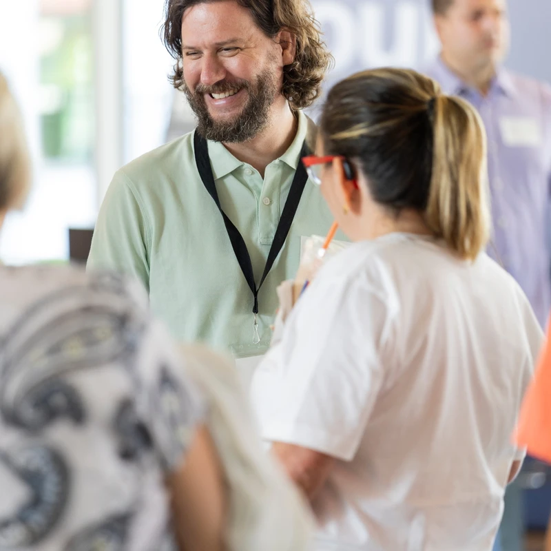 A man with long brown hair and a beard, wearing a green polo shirt and lanyard, smiles while talking with a woman at an indoor conference or event.