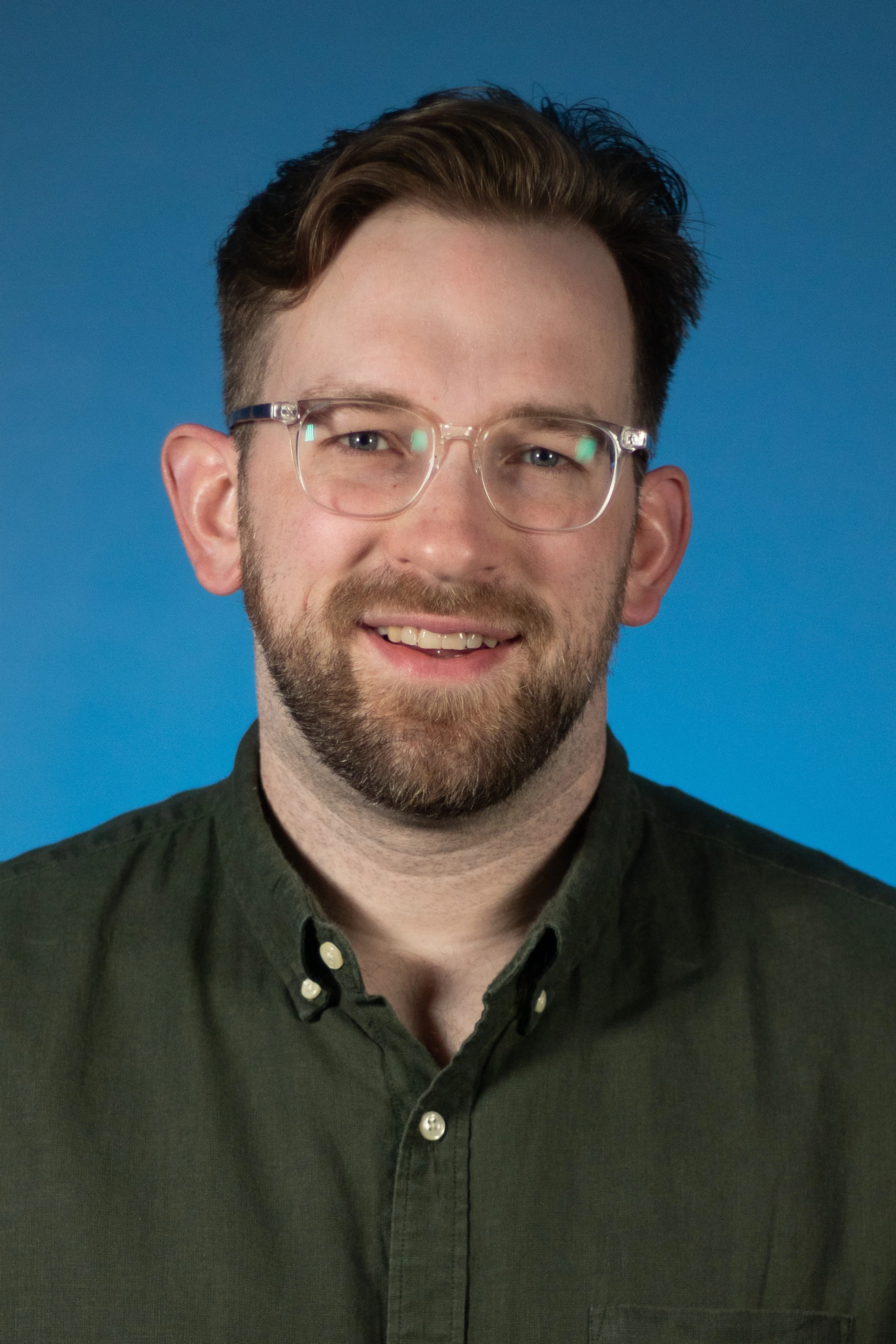 A headshot of a man with brown hair, a beard, and clear glasses, smiling at the camera. He is wearing a dark green shirt against a solid blue background.