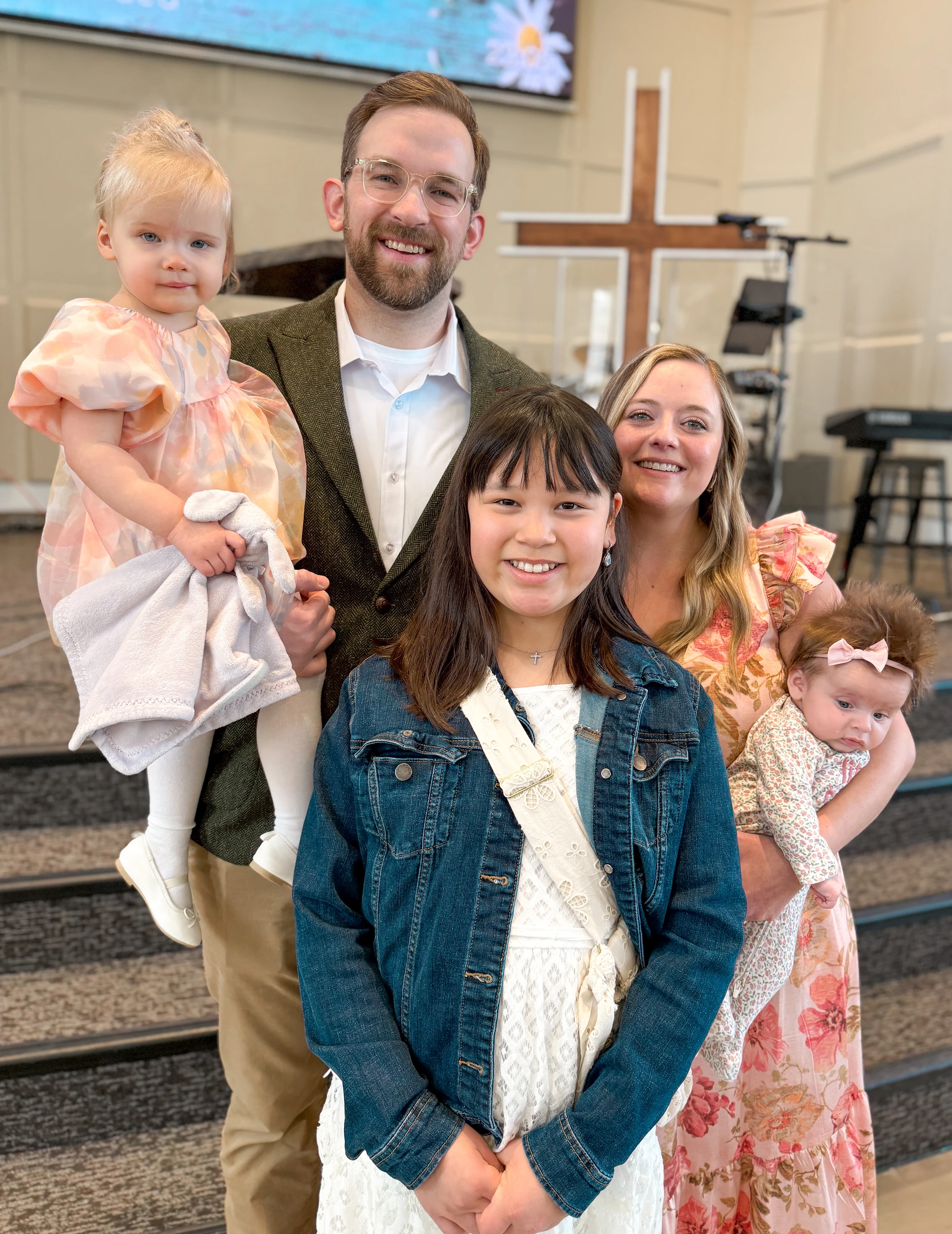 A smiling family of five, including a man, woman, older girl, toddler, and baby, pose together for a picture inside a church, with a wooden cross behind them.