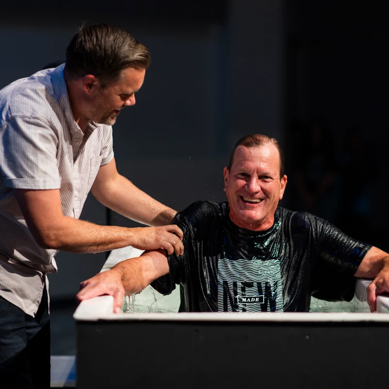 A smiling older man sits in a baptismal font, wet from the water, as a pastor stands beside him with hands on his shoulders during a baptism ceremony.