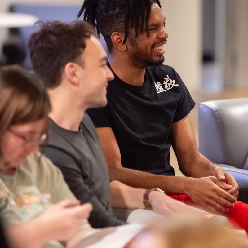 A young Black man with his hair in dreadlocks smiles broadly while sitting and listening in a group of people in a casual indoor setting.