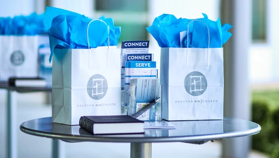 Two white welcome bags from Houston NW Church on a small table, next to a Bible and pamphlets titled 'Connect' and 'Serve' for new visitors.