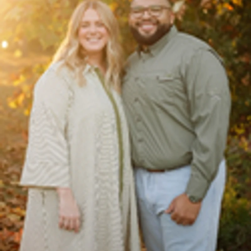 A smiling man and woman stand together outdoors in the warm glow of sunset, with fall foliage in the background.