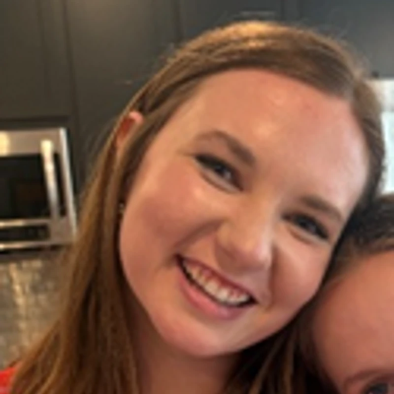 A close-up photo of a young woman with light brown hair smiling warmly at the camera. She is indoors with a kitchen in the background.