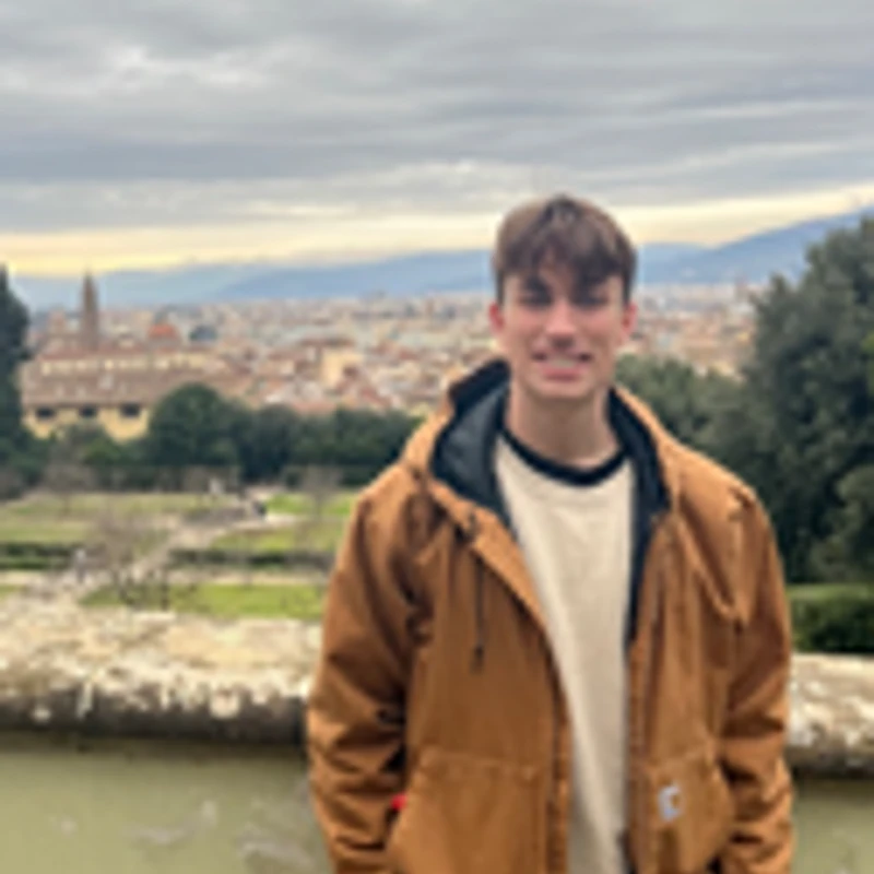 A young man in a brown jacket smiles for a photo. In the background is a scenic overlook of the city of Florence, Italy, under a cloudy sky.