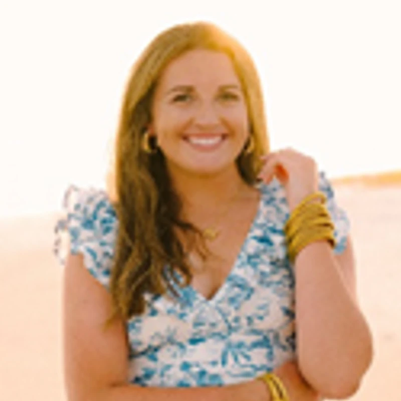A portrait of a smiling woman with brown hair wearing a blue and white floral dress and gold bracelets, standing outdoors during a warm sunset.